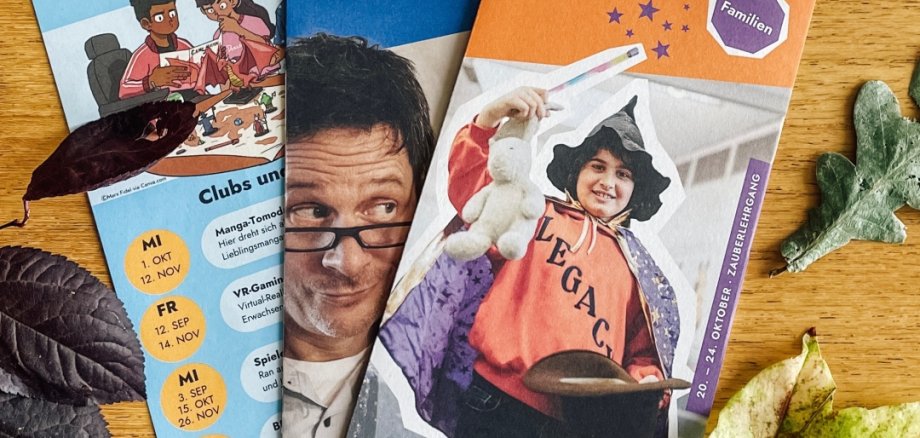 Auf einem Holztisch liegen drei Programmflyer ausgebreitet. Das rechte Programm trägt den Titel Familie: Programm Sep - Dez 2025 und ist orangefarben. In der Mitte des Flyers ist ein Kind mit Zauberhut und Zauberstab zu sehen. In der Mitte ist der Flyer blau und nur halb zu sehen. Hierbei handelt es sich um das Erwachsenenprogramm. Als Titelbild ist ein Mann mit Brille zu erkennen. Links außen ist der Flyer für das Jugend-Programm zu sehen. Oben auf dem Flyer ist eine Gruppe Jugendliche beim Pen-Paper Spielen zu sehen.