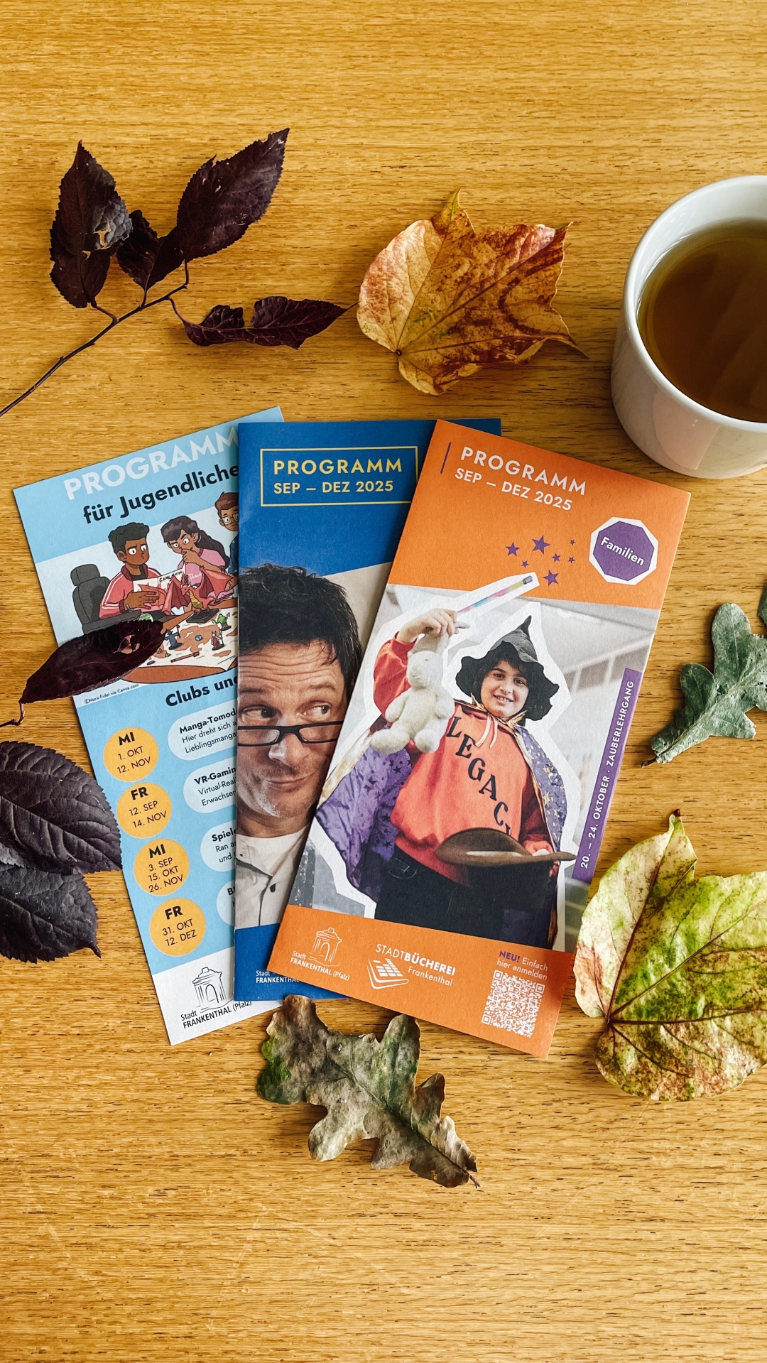 Auf einem Holztisch liegen drei Programmflyer ausgebreitet. Das rechte Programm trägt den Titel Familie: Programm Sep - Dez 2025 und ist orangefarben. In der Mitte des Flyers ist ein Kind mit Zauberhut und Zauberstab zu sehen. In der Mitte ist der Flyer blau und nur halb zu sehen. Hierbei handelt es sich um das Erwachsenenprogramm. Als Titelbild ist ein Mann mit Brille zu erkennen. Links außen ist der Flyer für das Jugend-Programm zu sehen. Oben auf dem Flyer ist eine Gruppe Jugendliche beim Pen-Paper Spielen zu sehen.