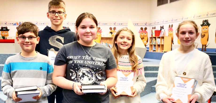 Fünf Kinder, zwei Jungs und drei Mädchen, stehen mit Büchern in der Hand vor dem Bilderbuchbereich in der Stadtbücherei Frankenthal und schauen lächelnd in die Kamera.
