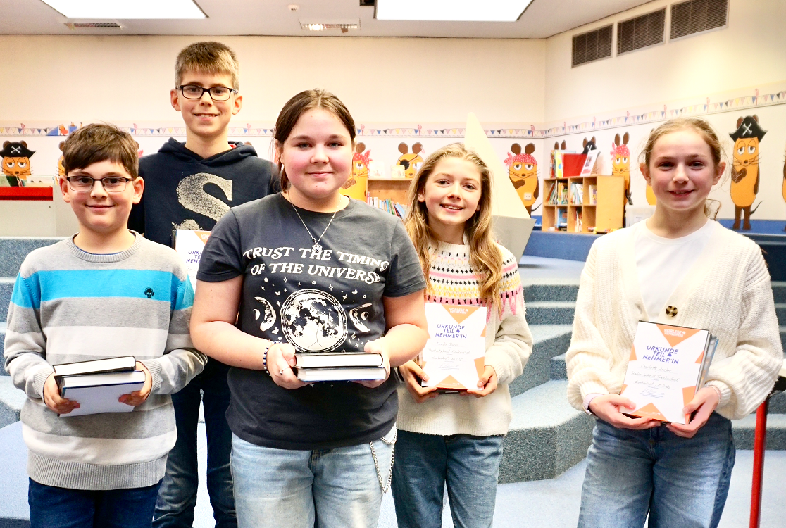 Fünf Kinder, zwei Jungs und drei Mädchen, stehen mit Büchern in der Hand vor dem Bilderbuchbereich in der Stadtbücherei Frankenthal und schauen lächelnd in die Kamera.
