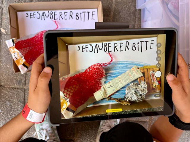 Mit Tablet Postkarten fotografieren Hände halten ein Tablet, mit dem gerade ein selbst gemaltes Bild fotografiert wird.