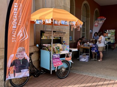 Lastenrad FRANKI auf der Kinderstadt 2026 Blick auf die orangene Fahne der Stadtbücherei Frankenthal und das hellblaue Lastenrad, auf dem viele Bücher ausgestellt sind. Im Hintergrund ein Tisch, an dem Menschen sitzen und stehen und malen.