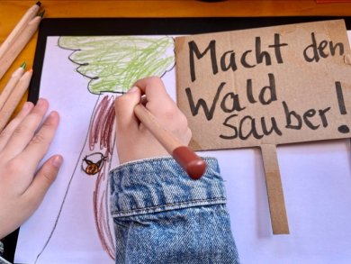 Bilder für eine saubere Umwelt malen Blick von oben auf ein weißes Blattpapier und Hände, die gerade einen Baum mit einem Buntstifte malen. Daneben liegt ein kleines Schild aus Pappe mit der Aufschrift: Macht den Wald sauber!