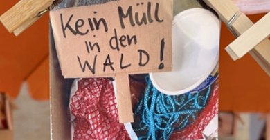Postkarten für eine saubere Umwelt Ein Foto hängt mit Holzklammern an einem Sonnenschirm. Auf dem Foto sieht man ein gemaltes Bild von einem Wald und einem Pappschild mit der Aufschrift: Kein Müll in den Wald!