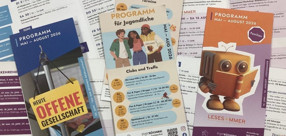 Auf dem Fotos sieht man die drei Faltblätter der Veranstaltungsprogramme Mai - August 2026 für Erwachsene, Jugendliche und Familien nebeneinander liegen. Im Hintergrund liegen weitere aufgeschlagene Faltblätter, ohne dass man einzelne Veranstaltungen genauer lesen kann.