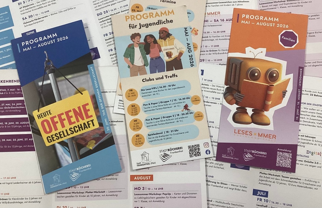 Auf dem Fotos sieht man die drei Faltblätter der Veranstaltungsprogramme Mai - August 2026 für Erwachsene, Jugendliche und Familien nebeneinander liegen. Im Hintergrund liegen weitere aufgeschlagene Faltblätter, ohne dass man einzelne Veranstaltungen genauer lesen kann.