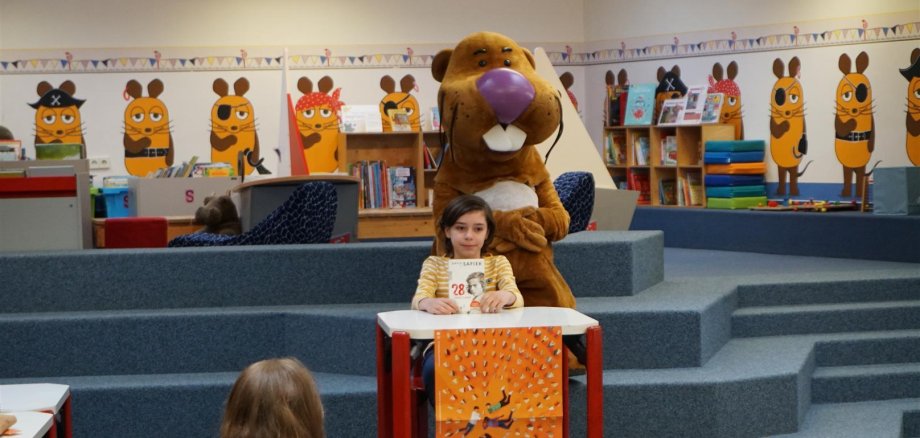 Ein Junge sitzt auf einer Bühne an einem Tisch und liest aus seinem Buch vor. Hinter ihm sitzt das Maskottchen der Rheinpfalz, der Bieber Nils Nager auf den Stufen zur Bilderbuchecke in der Stadtbücherei.