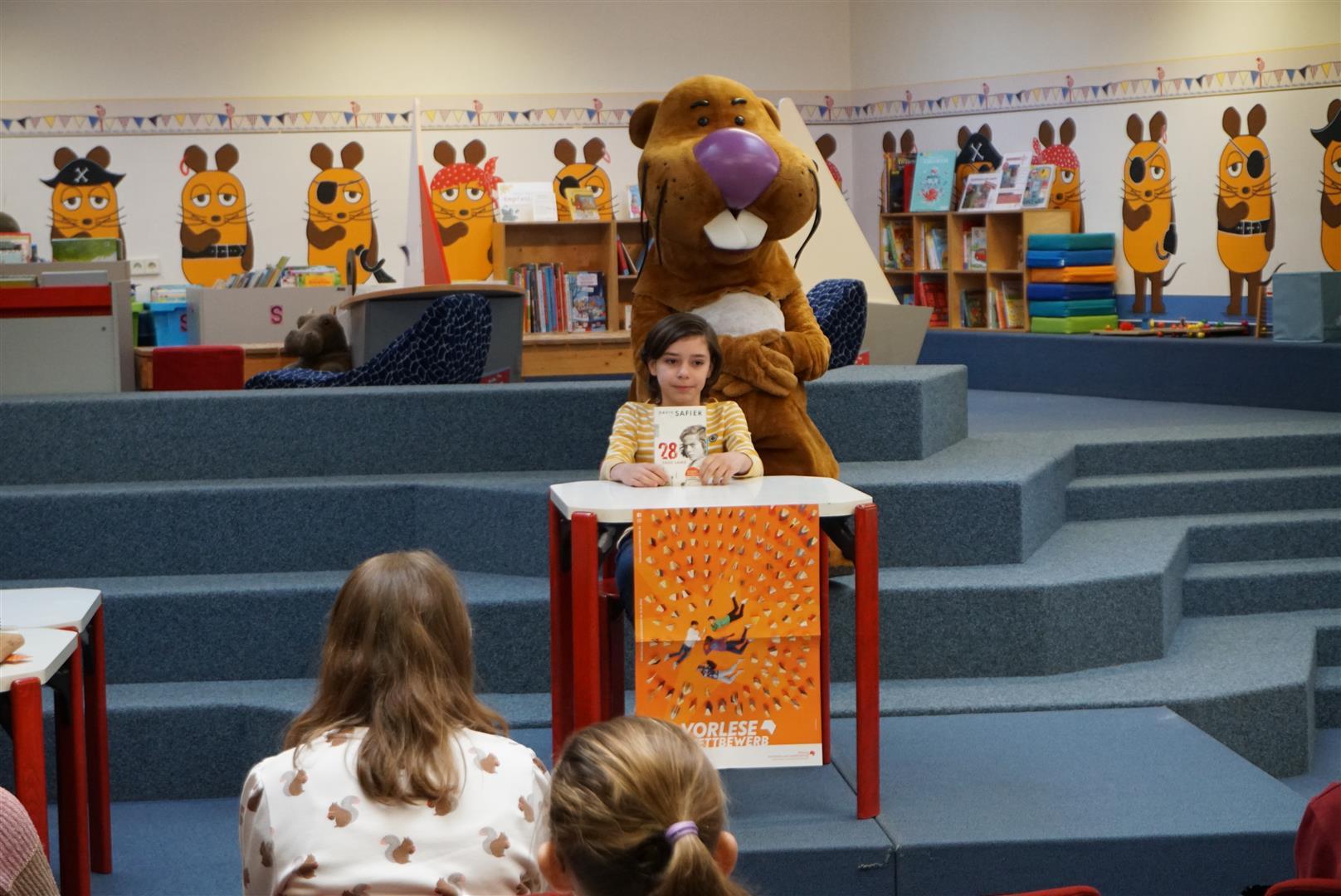 Ein Junge sitzt auf einer Bühne an einem Tisch und liest aus seinem Buch vor. Hinter ihm sitzt das Maskottchen der Rheinpfalz, der Bieber Nils Nager auf den Stufen zur Bilderbuchecke in der Stadtbücherei.
