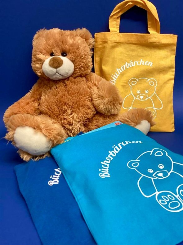 Das Bücherbärchen der Stadtbücherei Frankenthal Ein brauner Spielzeug-Teddybär sitzt neben bunten kleinen Stoffbeuteln vor dunkelblauem Hintergrund.