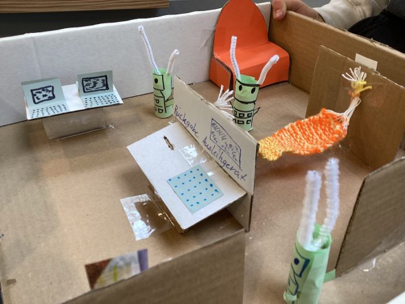 Aus der Vogelperspektive schaut man von oben auf das selbstgebaute Pappmodell einer Bücherei. Auf kleinen Papptischen stehen zwei kleine Papierlaptops und ein Rückgabeautomat. Zwischen zwei Wänden ist eine orangegelb gehäkelte Hängematte gespannt. In der hinteren rechten Ecke steht ein großes Sofa aus orangenem Papier. Drei Roboter aus grünen Klopapierrollen mit weißen Pfeifenputzerantennen stehen mittig im Modell.