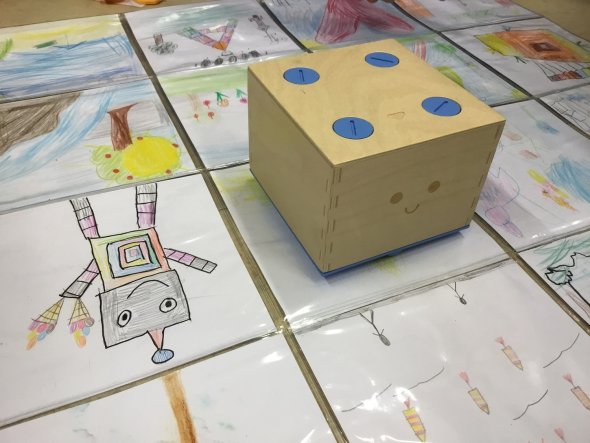 Cubetto unterwegs Der hölzerne Lernroboter Cubetto steht auf einer mit einem Roboter bemalten Boddenmatte.