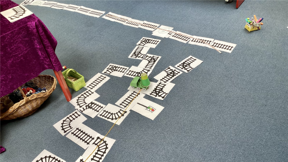 Messen mit Dr. Brumm Auf blauem Teppichboden liegen weiße Kärtchen, auf denen schwarze Schienen aufgemalt sind. So ergibt sich ein Netz aus mehreren Schienen verteilt über den Fußboden der Stadtbücherei.