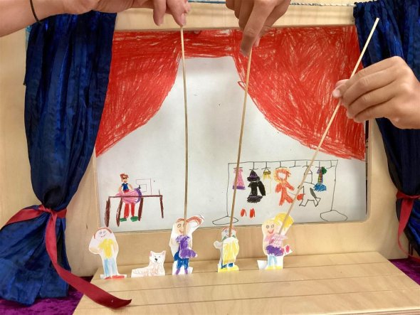 Vorhang auf! In einem Holzrahmen mit zur Seite gebundenem blauen Vorhang steht ein von einem Kind selbst gemaltes DINA3-großes Hintergrundbild. Davor werden vier Spielfiguren an Stäben mit einer Hand festgehalten.