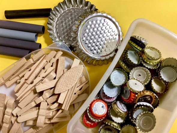 Klang-Werkstatt Aus der Vogelperspektive schaut man auf einen gelben Tisch mit Materialien zum Geräusche machen: hölzerne Klammern, metallene Flaschendeckel, graue Plastikrohre und silberne Tortenförmchen.