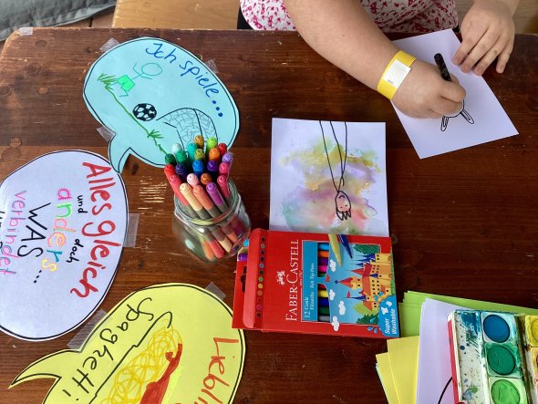 Auf einem braunen Holztisch sieht man von oben auf zwei Hände, die mit einem schwarzen Filzstift eine Strichzeichnung malen. Im Zentrum liegt ein weißes Papier mit einer schwarzen Strickzeichnung eines Männchens auf bunten Flecken aus Wasserfarben. Ein Glas mit Filzstiften, ein Kasten mit Wasserfarben und bunt beschriebene Sprechblasen sind auf dem Tisch verteilt.