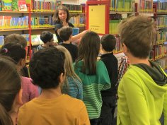 Bibliotheksführung in der Kinderbücherei Eine Mitarbeiterin steht vor einem Bücherregal führt eine Gruppe von Grundschülern durch die Kinderbücherei.