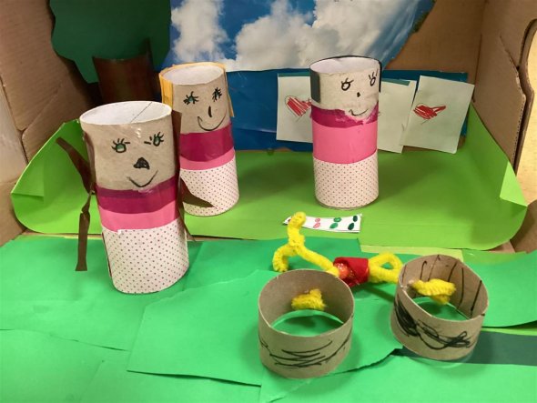 Man schaut in einen Schuhkarton wie auf eine Bühne. Auf Rasen aus grünem Papier stehen drei bemalte und rosa beklebte Figuren aus Papprollen und Papier.