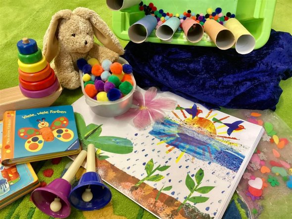 Veranstaltungen für die Kleinsten bis 3 Jahre Auf grünem Hintergrund liegen verschiedenste Materialien, wie Bilderbücher, Glöckchen, Papprollen, ein Kuschelhase, Holzspielzeug oder Stoffkügelchen.
